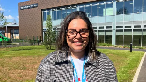 Photograph of Ruth Elliott from Royal College Manchester. The image shows her in front of the newly built Ged Mason building. She has shoulder-length dark hair and wears glasses. Smiling, she is wearing a checked jacket on top of a white top.