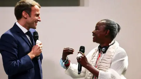AFP/Getty Images Kouoh speaks to a besuited Macron, who smiles back at her. Both are holding microphones.