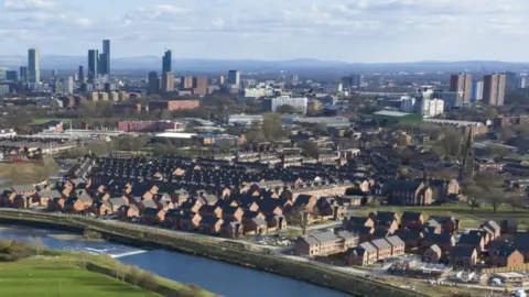 The Greater Manchester skyline, with tower blocks and skyscrapers in the background and an estate of brick-built houses in the foreground