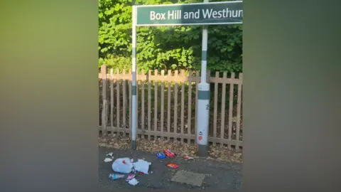 LDRS Litter including food packaging, bottles and bags is scattered on a platform beneath a Box Hill & Westhumble station sign on 5 July 2026, with trees and fencing in the background.