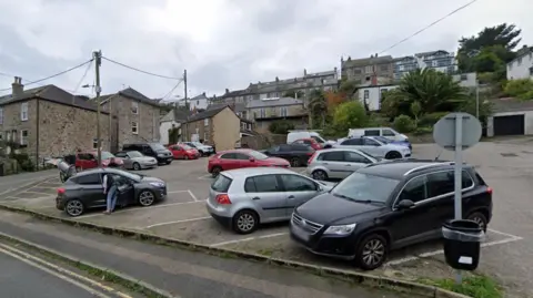 A small car park which is nearly full, with one person seen looking into the back seat of their vehicle