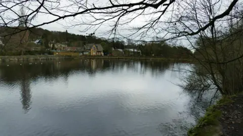 A large patch of water with overhanging trees and vegetation, At the far end of the water there are a collection of buildings.