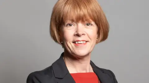 A woman with short brown hair wearing a black suit jacket and red top smiles as she poses for the camera