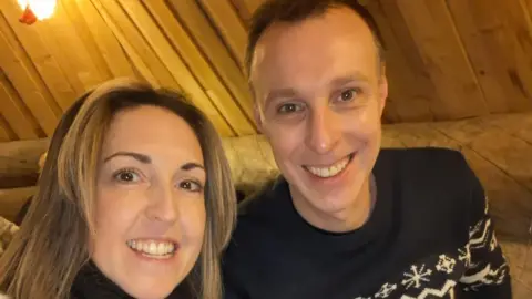 David Eddon A man and a woman sitting inside a wooden cabin. They are shown from the shoulders up and are both smiling at the camera. The woman, on the right has brown shoulder length hair and is wearing a black polo neck jumper and the man has short brown hair and is wearing a nordic-style black and white jumper. 