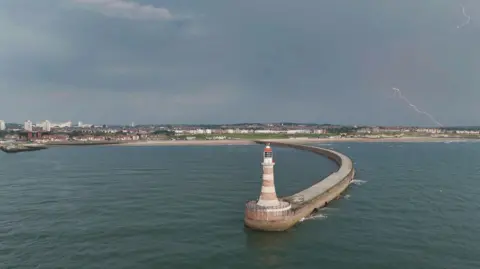 Richard R Clark A curved pier with a small striped lighthouse at the end of it. The sea around it is dark and calm. Two faint bolts of lightning can be seen on the right, hovering over housing on the shoreline.