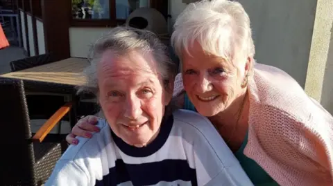 Merseyside Police Eric Greener, who has grey hair swept to the side and is wearing a blue and white striped sweatshirt, smiles at the camera while sitting in a chair outside a pub or restaurant. Sheila Jackson, who has short white hair and is wearing  pink cardigan leans into the frame and smiles at the camera with her arm around Greener's shoulder.