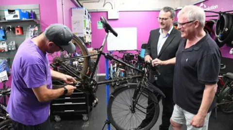 A bike is up on a stand, and the mechanic is doing something to the chain, while Mark and Fergal look on.  There are bikes and tyres in the background and the walls are painted a bright purple/pink.