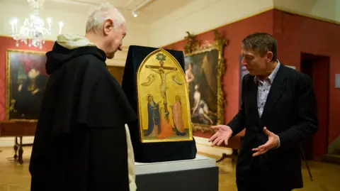 Ian Wallman Dr Xa Sturgis at the press call announcing the purchase of the painting. Dominican Friar Timothy Radcliffe, OP is also in attendance. They are standing in front of the painting. There are other paintings behind them. 