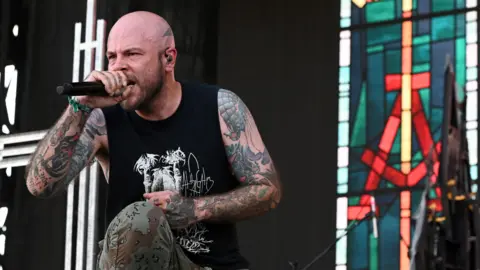 Getty Images Picture of Ryan Clark, who is the lead singer of metal band Demon Hunter. He is crouched on stage wearing a black vest and camouflage trousers and is holding a microphone.