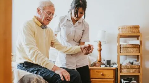 Getty Images An older man with a younger nurse, who is holding his left hand. Some bedroom furniture is pictured to the left of him.