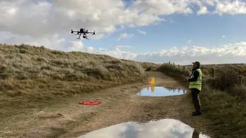 Natural England A drone flies in the air at a low level over a stretch of fenced off dune, with a male operator - dressed in a high-vis vest and baseball hat - standing nearby on a path covered in puddles and with the drone control in his hands.