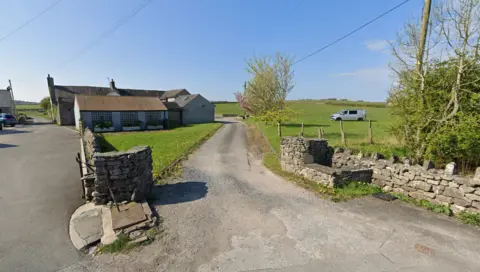 Road leading up to Nook Farm Bungalow in Ulverston. The farm sits in a green field with cottages to its left. It is a sunny day.