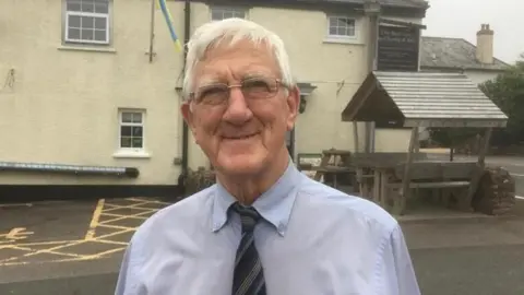 Eric Norman, a man in his 70s wearing glasses, a blue shirt and striped tie, is stood in a pub car park with the building behind him