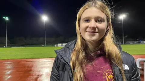 Maddie Thomson is pictured standing in front of a flood-lit running track on a rainy night. She has long, blonde hair and is smiling. She is wearing a burgundy sweatshirt and a black North Face puffer jacket.