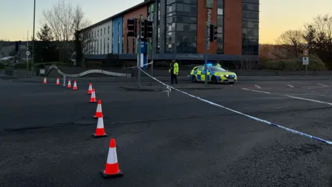 Danny Fullbrook/BBC A road closed with cones across the road, police tape and an officer with a car.