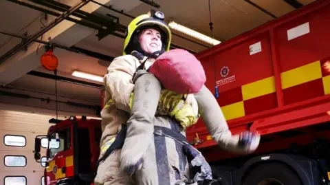 Emma Ward on duty as a firefighter, carrying a dummy, with a fire engine parked behind her.