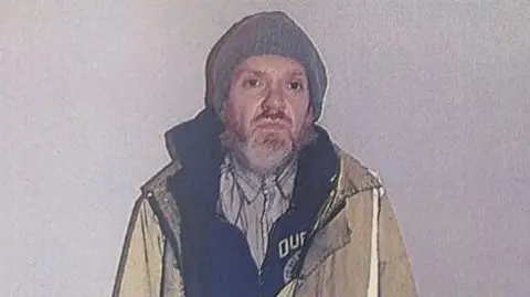 Roy Barclay, a bearded man with brown eyes, is in a yellow hi-vis jacket. He is also wearing a grey beanie, a striped shirt, and a dark hoodie with a logo or writing which is not fully visible behind his jacket.