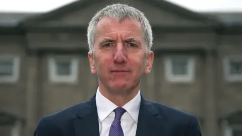 Archive image showing Máirtín Ó Muilleoir with short grey hair, wearing a suit, in front of a building.