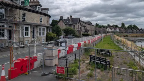 The footpath by the river on Aynam Road in Kendal is closed. There are white and red road blocks and fencing along the length of it.