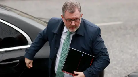 Sir Jeffrey Donaldson arrives at Newry Crown Court. He wears a blue suit, white shirt and green tie. He has grey hair and wears glasses. There is a black car behind him. 