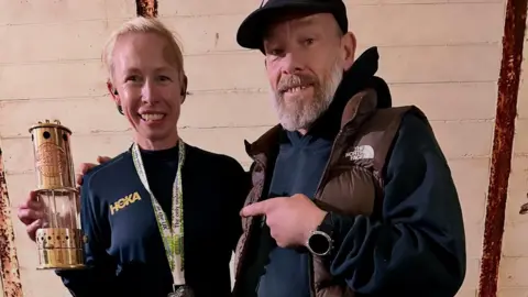 Laura Watts A smiling Laura Watts with Jay McCardle. Laura, who has blonde hair, is holding up her marathon trophy, a miners oil lamp.