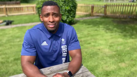 BBC Olympian Joel Fearon is sitting at a table in a garden and is wearing a blue Great Britain t-shirt with the Olympic rings on it.