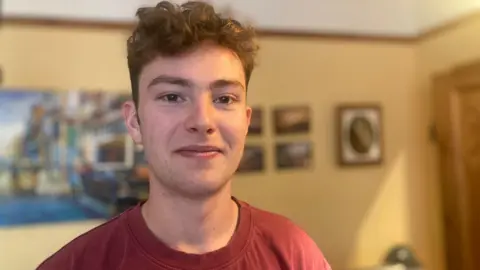 Mousumi Bakshi/BBC Manolo Niembro is smiling at the camera while wearing a red t-shirt. He has  brown eyes and light brown hair that is short on the sides and curly on top.  He is stood in a room with creamy yellow walls and framed pictures are behind him. 