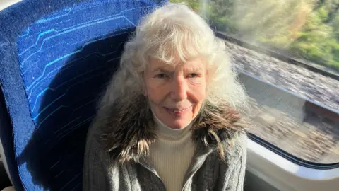 BBC/RICHARD EDWARDS Big Communi-Tea client Ann Waddington on the train to London. She is sitting in a window seat, smiling, and is wearing a white polo neck and a grey cardigan with a fur-trimmed collar.