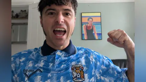 James Baker pictured in a 1992-94 Coventry City shirt. It is blue and white with the club's crest on his left breast and sponsor's name Peugeot. He is standing in a room with furniture and a painting on the wall. 