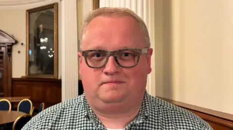 Séamas de Faoite has short grey hair, glasses and a checked shirt. 