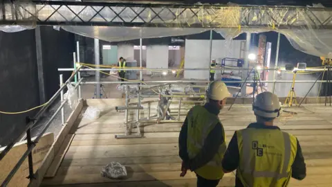 Two men in white hard hats and florescent jackets on a light wood scaffold looking down towards three more colleagues below.