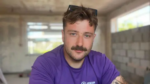 A man with brown hair, wearing sunglasses on his head and in a purple t-shirt on a building site.
