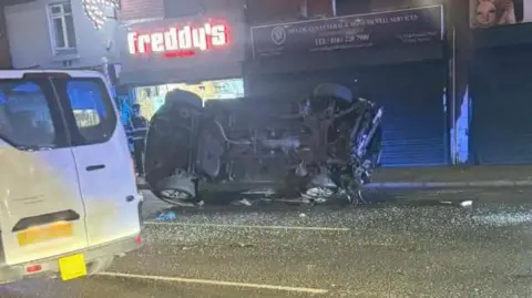 Handout An overturned car lies on its side on Hyde Road, Gorton. Only its underside is visible. Shattered glass can be seen in the road next to it. A white taxi is to its left and shops behind it.