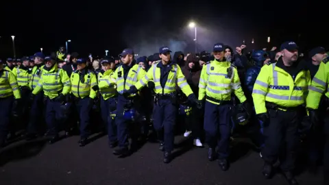 Getty Images A line of more than a dozen police officers in high-visibility jackets and police hats. They are linking arms in front of a large crowd of football supporters, all of whom are wearing dark clothing. 