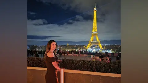 Collect/PA Real Life A woman with long brown hair is wearing a brown dress and is smiling as she stands on a balcony that is looking out towards the Eiffel Tower in Paris. The tower is lit up because it is night time, and it is a golden light with white lights below it.