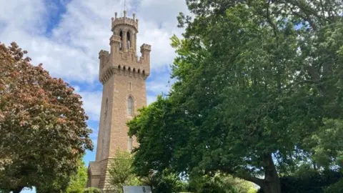BBC Guernsey's Victoria Tower is seen through the trees while the sky in the background is blue with a lot of cloud coverage 