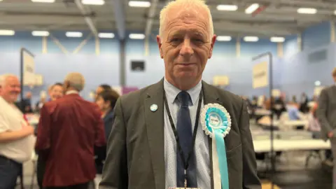 A man wearing a blue Reform UK badge smiling into the camera