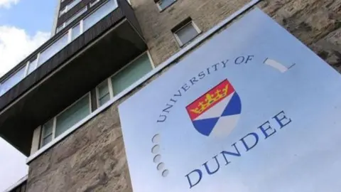 A sign reads 'University of Dundee' around a crest. The sign hangs on a building and the camera is angled up towards the ascending storeys.