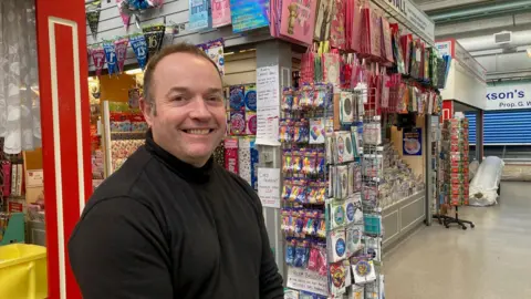 A man in a black polo neck standing in front a card shop 