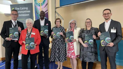 Essex County Council A group of seven smartly dressed men and women holding copies of the report. They are inside a conference room. The carpet is blue and there is some wooden furniture behind them.