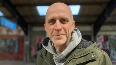 Mark Deans is the chairman of the Flo Charitable Trust. He is looking straight towards the camera and is wearing a grey hooded top, with a green coat on top. 