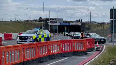Helen Mulroy/BBC A police truck has its blue lights on behind a line of orange barriers shutting the road. It is blocking the entrance to a tunnel at the airport, which has a stream of cars queuing to get out.