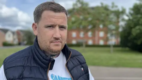 Vinnie Condron standing in a park with trees, grass and a red brick six-story building in the distance. He has short cropped brown hair and a light beard that is reddish on his chin. He has small neck tattoo. He's wearing a puffy navy blue gilet and a white long sleeve cotton shirt. 