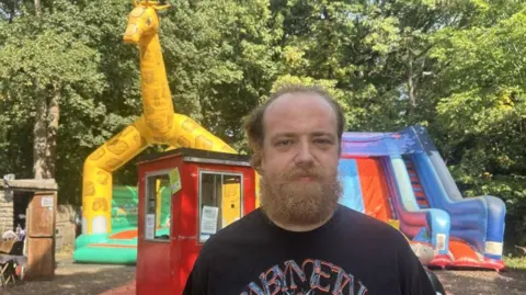 A man with light brown hair and a fuzzy beard is standing in the foreground. He is wearing a black t-shirt with writing on it. Behind him there is a bouncy castle with 