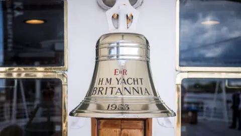 Getty Images A close-up of a polished metal ship’s bell mounted on a white wall. The bell is engraved with the words “H.M. Yacht Britannia” and the year “1953,” with windows on either side.