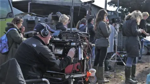 Wellington Films A film crew and camera man sat behind a large camera and filming equipment. There are lots of people standing around in coats and wellies on a muddy patch of ground, wearing headphones and facing in the direction of the actors.