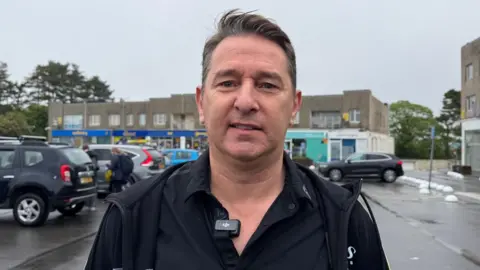 Joe Pinzari is wearing a black jacket and black T-shirt. He is pictured in front of Les Quennevais Parade. 