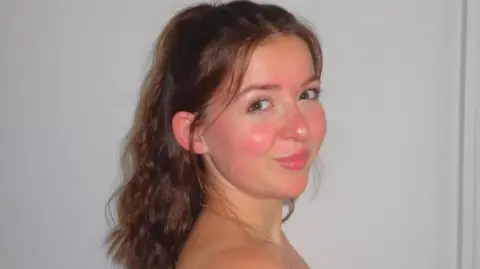 Mia Mia. She has long dark hair which is half tied up. She is looking over her shoulder at the camera and smiling.