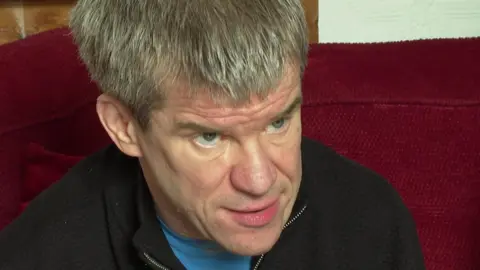 Darren Hanley, who has grey thick hair and blue eyes, wears a blue t-shirt and a black jumper with the zip opened.