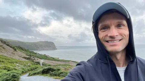 Hans Obma Hans Obma smiles directly at the camera while holding it in his left hand. He stands in the right of the frame and wears a hooded jacket over a hat. Behind him, the coast of Llyn Peninsula
